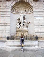 young attractive happy sunny girl in gray pants, cloak and sunglasses is standing fashion with gold backpack and posing on Trieste sculpture background in Italy and smiling