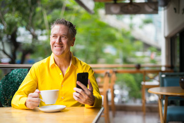 Mature Man Sitting In Coffee Shop And Thinking While Using Mobile Phone