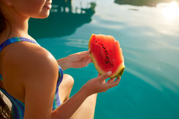 Pretty woman holds slice red watermelon stretching long tanned legs over blue pool, relaxing on...