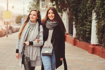 two young happy girlfriends walking on city streets in casual fashion outfits, wearing warm coats and having fun