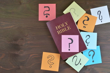 Holy bible and colorful note pads with question marks on brown wooden background as symbol for...