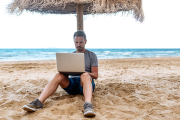 man leaning on a straw umbrella at the beach with laptop 