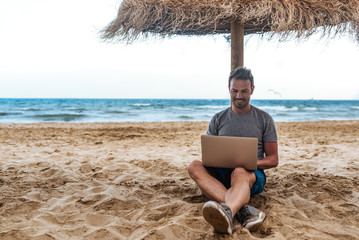man leaning on a straw umbrella at the beach with laptop 