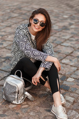 Fashionable happy girl in a stylish jacket with a pattern with a bag sitting on a stone road