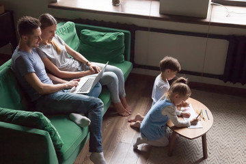 Kids son daughter drawing while parents using laptop at home on weekend, children siblings playing together having fun with family mom and dad entertaining with computer spending time in living room