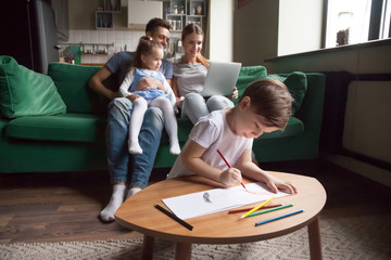 Creative kid boy son playing drawing with colored pencils while parents with little girl sister using laptop on sofa in living room, family children hobbies and activities at home on weekend concept