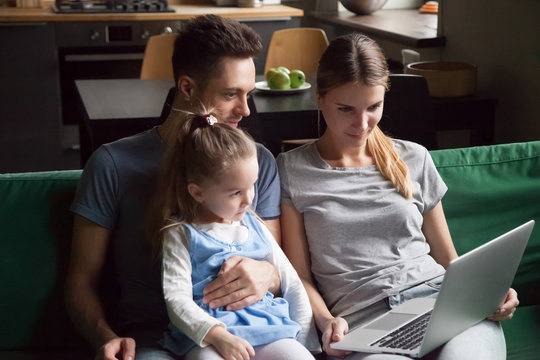 Young family with child daughter looking at laptop screen watching video together on sofa at home, parents and kid girl relaxing using computer application for shopping or education on weekend