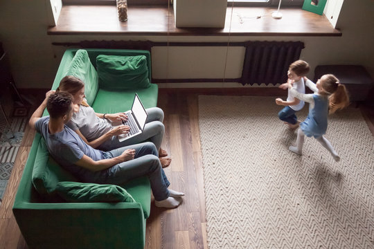 Happy Active Kids Running In Living Room Playing Game Having Fun Together While Parents Relaxing On Sofa Using Laptop, Family With Children Weekend Activities At Home Concept, Overhead Top View