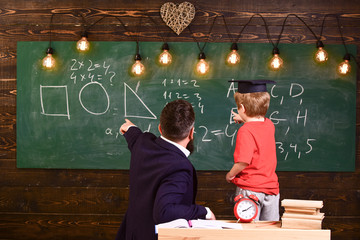 Teacher with beard, father teaches little son in classroom, chalkboard on background. Basics of education concept. Boy, child in graduate cap looks at scribbles on chalkboard while teacher explains.