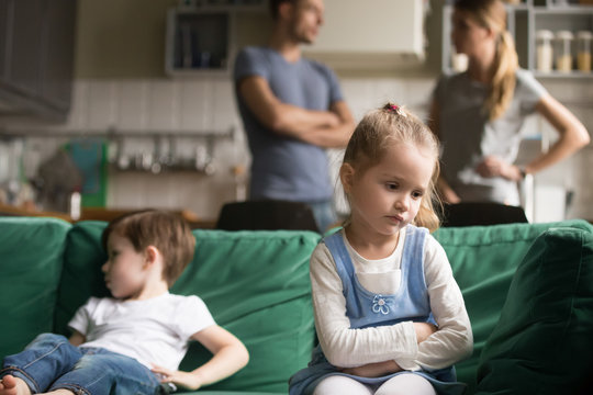 Upset little girl feeling sad after fight with brother sitting on sofa with worried parents on background, sulky frustrated sister ignoring child boy disinterested or bored, siblings rivalry concept