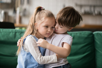 Little boy hugging consoling upset girl sitting on sofa, kid brother embracing sad sister...