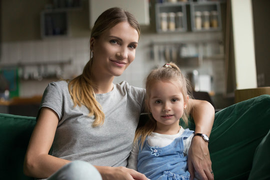 Portrait of loving happy family single mother embracing cute little girl sitting on sofa, smiling mom hugging preschool child daughter looking at camera at home, young woman and kid posing indoors