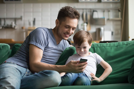 Smiling dad with kid son watching funny video on mobile phone at home, young father teaching little boy to use applications or play game on smartphone enjoying spending time online together on sofa