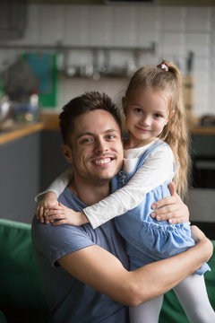 Vertical portrait of happy kid daughter embracing father at home, cute preschool girl hugging her smiling dad bonding looking at camera, caring daddy and little child good relationships concept