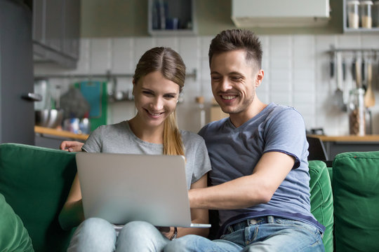 Happy millennial couple laughing looking at laptop screen together enjoying online humor, smiling young man and woman having fun at home on sofa using computer watching funny video in social networks