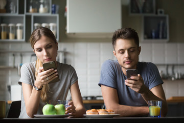 Millennial couple obsessed with smartphones ignoring each other having breakfast together at home, serious young man and woman using mobile apps or checking social networks online, gadgets overuse