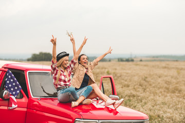 beautiful young girlfriends sitting on car hood with raised hands and showing peace signs © LIGHTFIELD STUDIOS