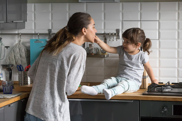 Little daughter feeding mother having fun in the kitchen at home, cute girl helping young single...