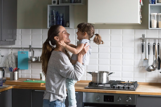 Smiling Loving Single Mother Holding Cute Little Child Daughter Having Fun Together, Happy Family Of Young Caring Mom And Excited Kid Girl Laughing Playing At Home While Cooking In The Kitchen