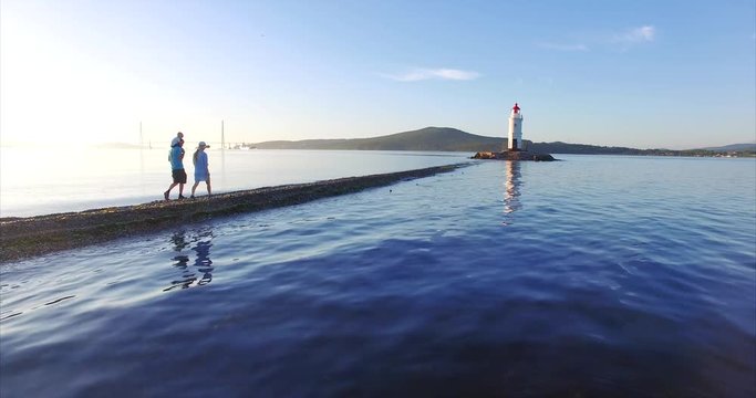 Beautiful view of family walking along the spit towards Tokarevsky lighthouse: father is holding his son on his shoulders. Morning, calm blue sea. Vladivostok. Russian bridge is on the background