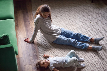 Cute kid girl lying on warm floor relaxing spending time with sister, young mom or babysitter at home, mother and child girl resting talking after playing together in living room on weekend