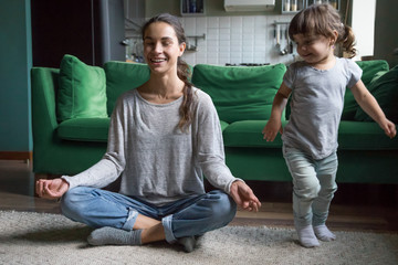 Happy mindful single mother doing morning exercises in yoga pose with kid daughter playing at home,...