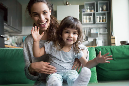 Portrait of happy excited young mother and kid girl waving hands looking at camera, smiling mom with child daughter making video call, family vloggers recording video blog or vlog together concept