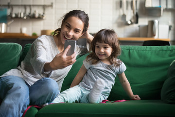 Happy mother laughing taking selfie with child daughter on smartphone at home, smiling single mom...