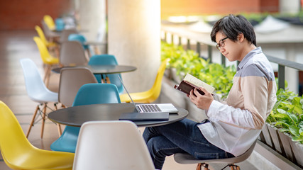 Young Asian student man with eyeglasses dressed in casual style reading book and using laptop computer in college building. Education research and self studying in university life concepts