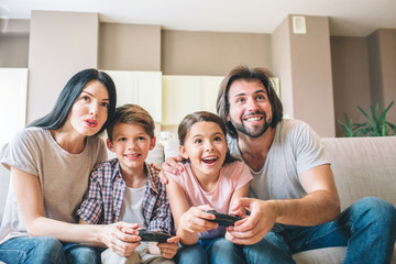 Excited people are sitting together on sofa. Parents help to play their kids. Children are amazed. look straight forward with their mouths opened.