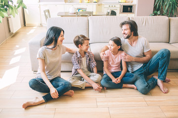 Positive and attractive people are sitting on the floor. They look at each other. Parents and kids wear jeans and shirts. They sit with legs crossed.