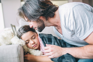 Girl has fallen asleep. She is covered with blanket. Her dad is sitting besides her. He is going to kiss child.