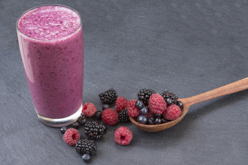 A lot of raspberries, blackberries and blueberries scatter from a wooden spoon and smoothies from these berries on a black background.