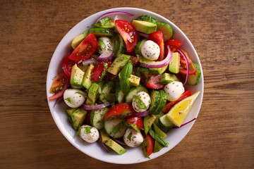 Avocado, tomato, cucumber salad with mozzarella cheese in white bowl on wooden table. overhead, horizontal