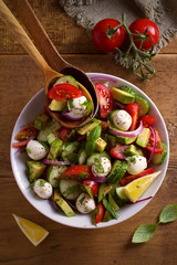 Avocado, tomato, cucumber salad with mozzarella cheese in white bowl on wooden table. overhead, vertical