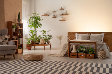 Warm bedroom interior with plants, shelves, striped rug, pouf, bed and armchair. Real photo. Place your graphic