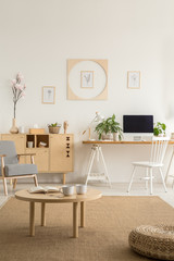 Wooden table and pouf on brown carpet in white workspace interior with posters and armchair. Real photo