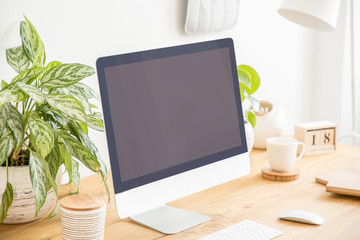 Mockup of black computer desktop on wooden desk with plants in white workspace interior. Real photo