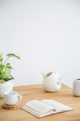 Book, cup and plant on wooden table against white wall in simple living room interior. Real photo