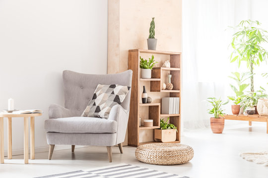 Real photo of a bright living room interior with an armchair, pillow, pouf and modern shelf