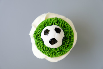 Chocolate cupcake on a football theme with white whipped cream decorated with green grass and soccer ball on gray background. Top view.