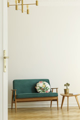 Floral pattern pillow on a retro style, green sofa and a wooden table in a minimalist living room interior with white wall and herringbone floor. Real photo.