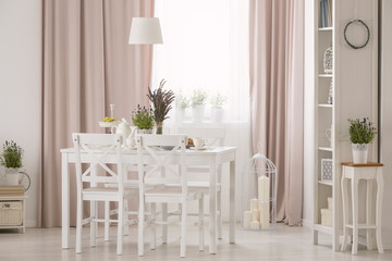 Lamp above table and white chairs in pink dining room interior with plants and drapes. Real photo