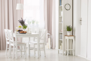 White chairs at dining table with plants in modern flat interior with pink drapes and window. Real...