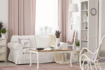 Wooden table next to white couch in pink living room interior with drapes and plants. Real photo