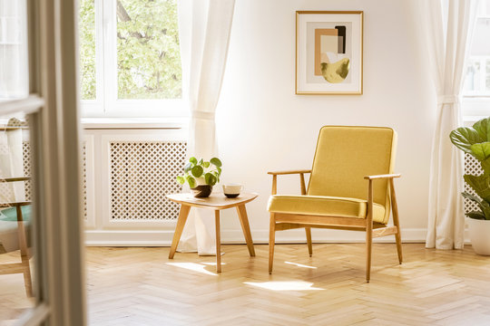 A retro, yellow armchair and a wooden table in a beautiful, sunny living room interior with herringbone floor and white walls. Real photo.
