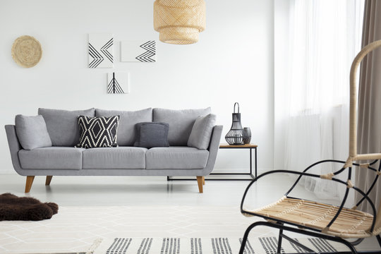 Real photo of a rattan armchair opposite a gray settee in scandi living room interior with geometric paintings on white wall