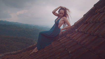 Aerial drone view of beautiful woman in long blue dress sitting on tiled red roof of the house against amazing mountain view and cloudy sky background - obrazy, fototapety, plakaty