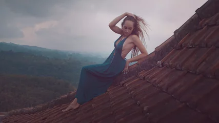 Foto op Aluminium Aerial drone view of beautiful woman in long blue dress sitting on tiled red roof of the house against amazing mountain view and cloudy sky background © stryjek