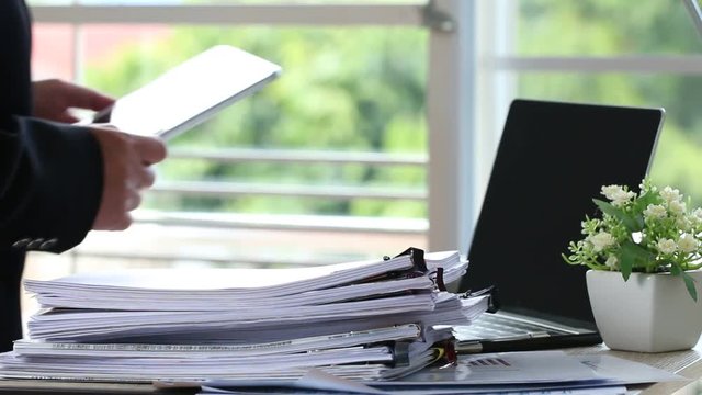 Stack unfinished documents paper report files and laptop on busy overwork desk with blur start up businessman working using apps for searching information in tablet. Business concept, vintage tone